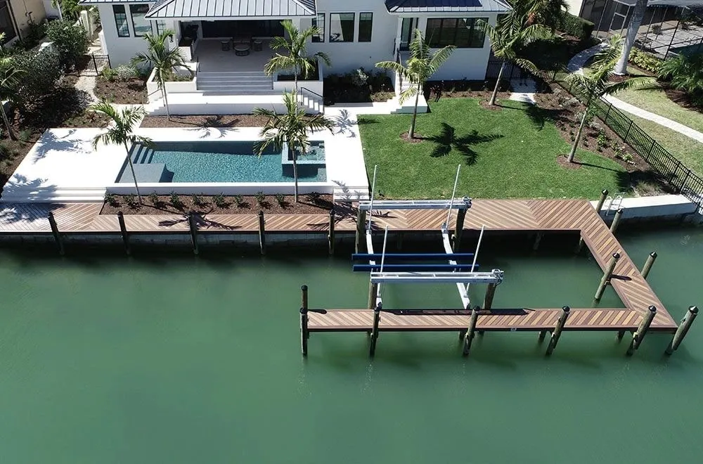 Aerial view of a wraparound hardwood dock with boat lift tracing the seawall of a modern waterfront home with a pool